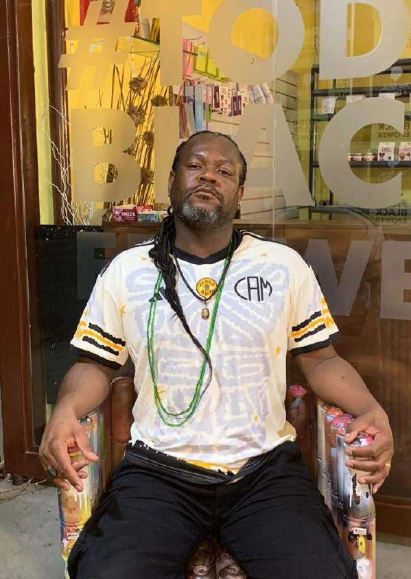 Fred Maciel, Negro F., sentado em frente a uma vitrine grafitada, vestindo camisa branca com grafismos e correntes africanas
